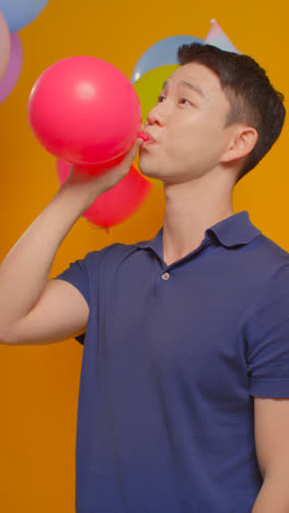 Retrato-De-Estudio-De-Vídeo-Vertical-De-Un-Hombre-Vestido-Con-Sombrero-De-Fiesta-Celebrando-Un-Cumpleaños-Inflando-Un-Globo